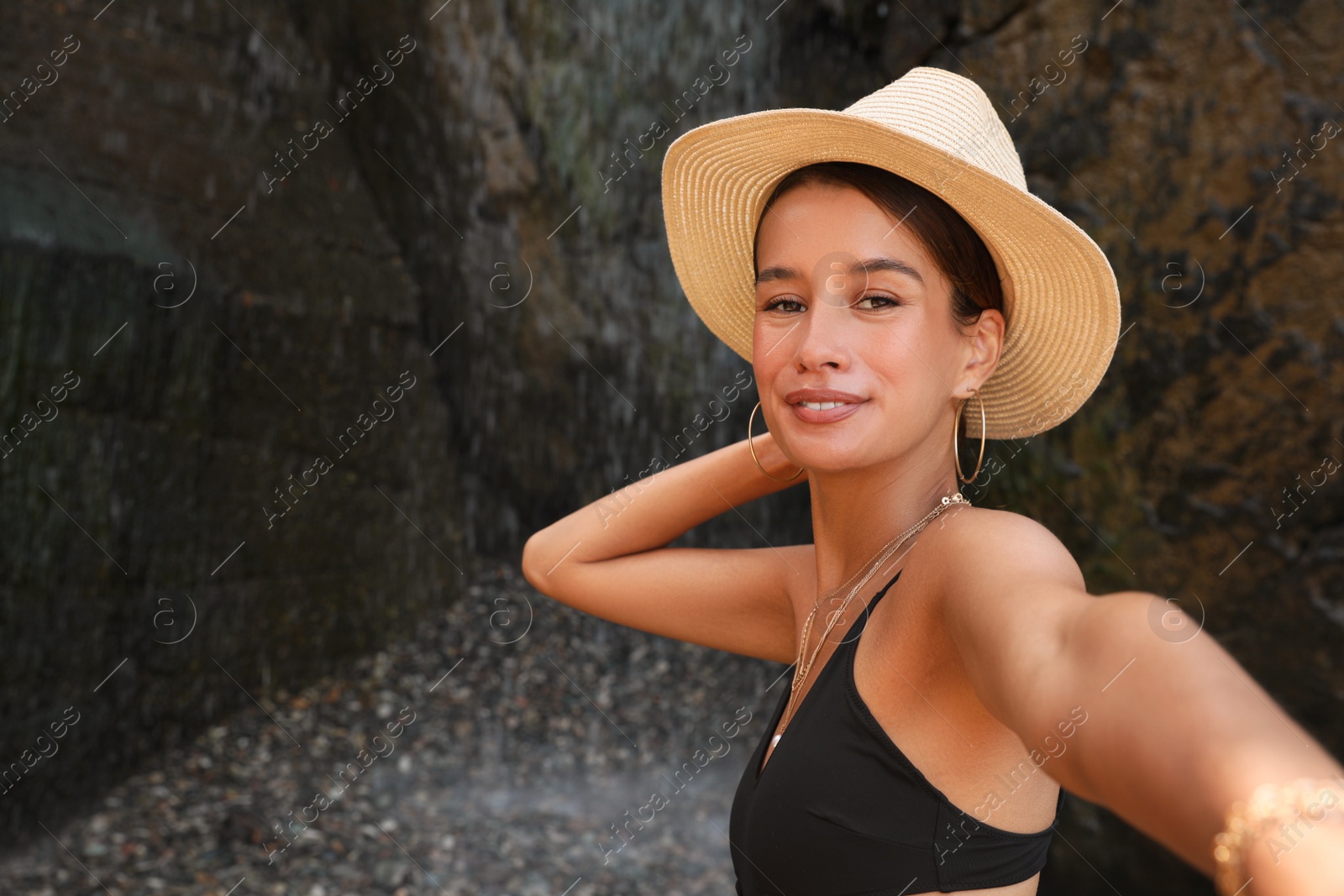 Photo of Beautiful young woman in stylish bikini taking selfie near waterfall outdoors, space for text