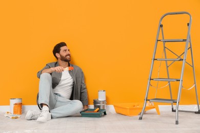 Photo of Happy designer sitting on floor near freshly painted orange wall indoors