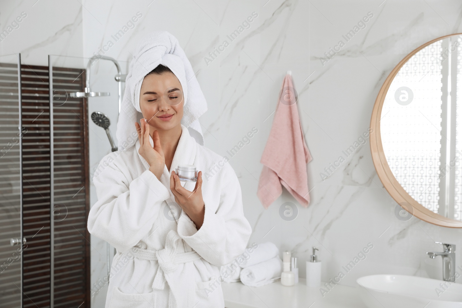 Photo of Beautiful young woman with hair wrapped in towel applying cream indoors