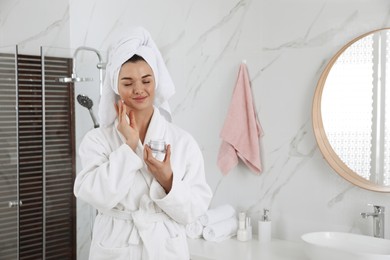 Photo of Beautiful young woman with hair wrapped in towel applying cream indoors