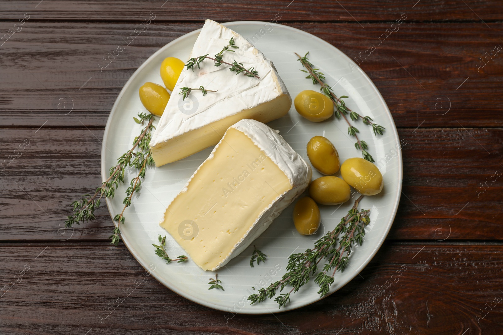 Photo of Plate with pieces of tasty camembert cheese, olives and rosemary on wooden table, top view