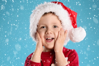 Cute child in Santa hat under snowfall on light blue background. Christmas celebration