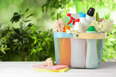 Photo of Plastic basket with spring flowers and cleaning supplies on white wooden table. Space for text