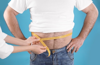 Doctor measuring overweight man's waist on color background, closeup