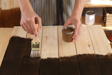 Man with brush and can applying wood stain onto wooden surface indoors, closeup