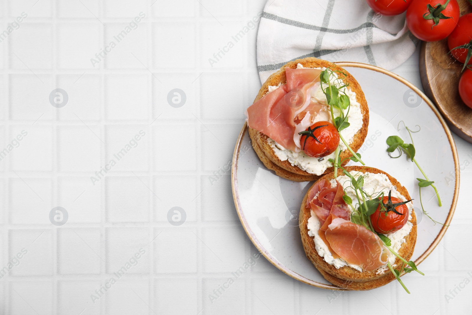 Photo of Tasty rusks with prosciutto, cream cheese and tomatoes served on white table, flat lay. Space for text