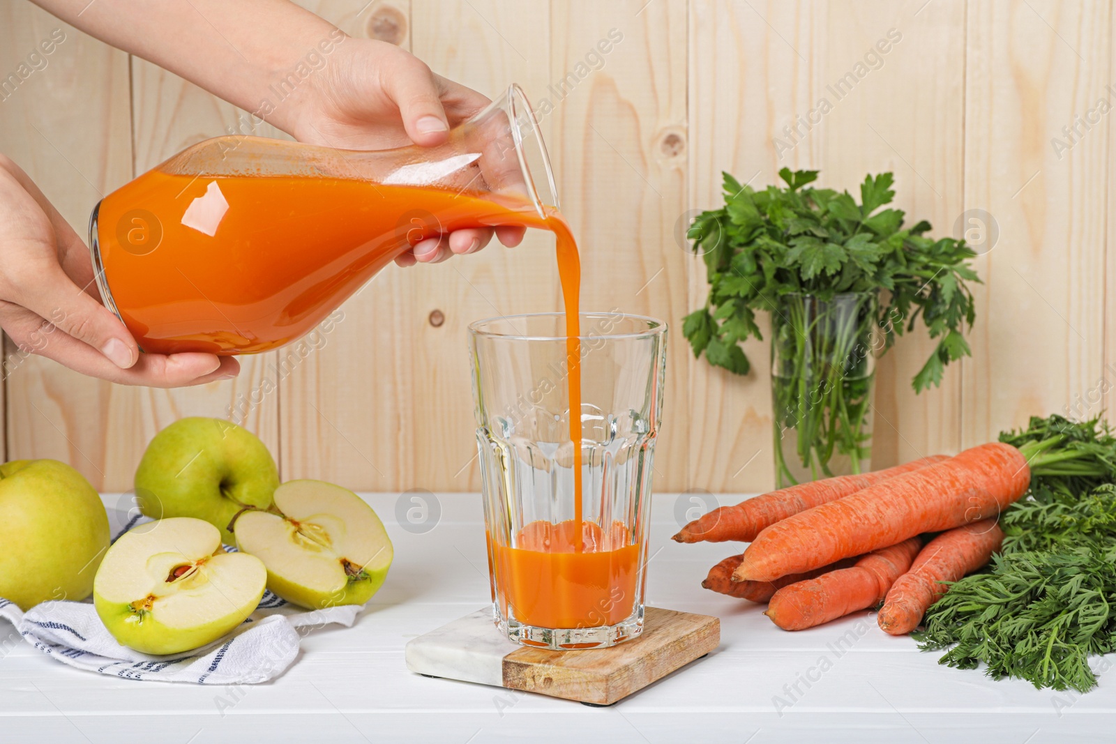 Photo of Woman pouring carrot juice from jug into glass at white wooden table, closeup