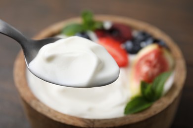 Spoon with tasty yogurt above bowl on table, closeup. Space for text