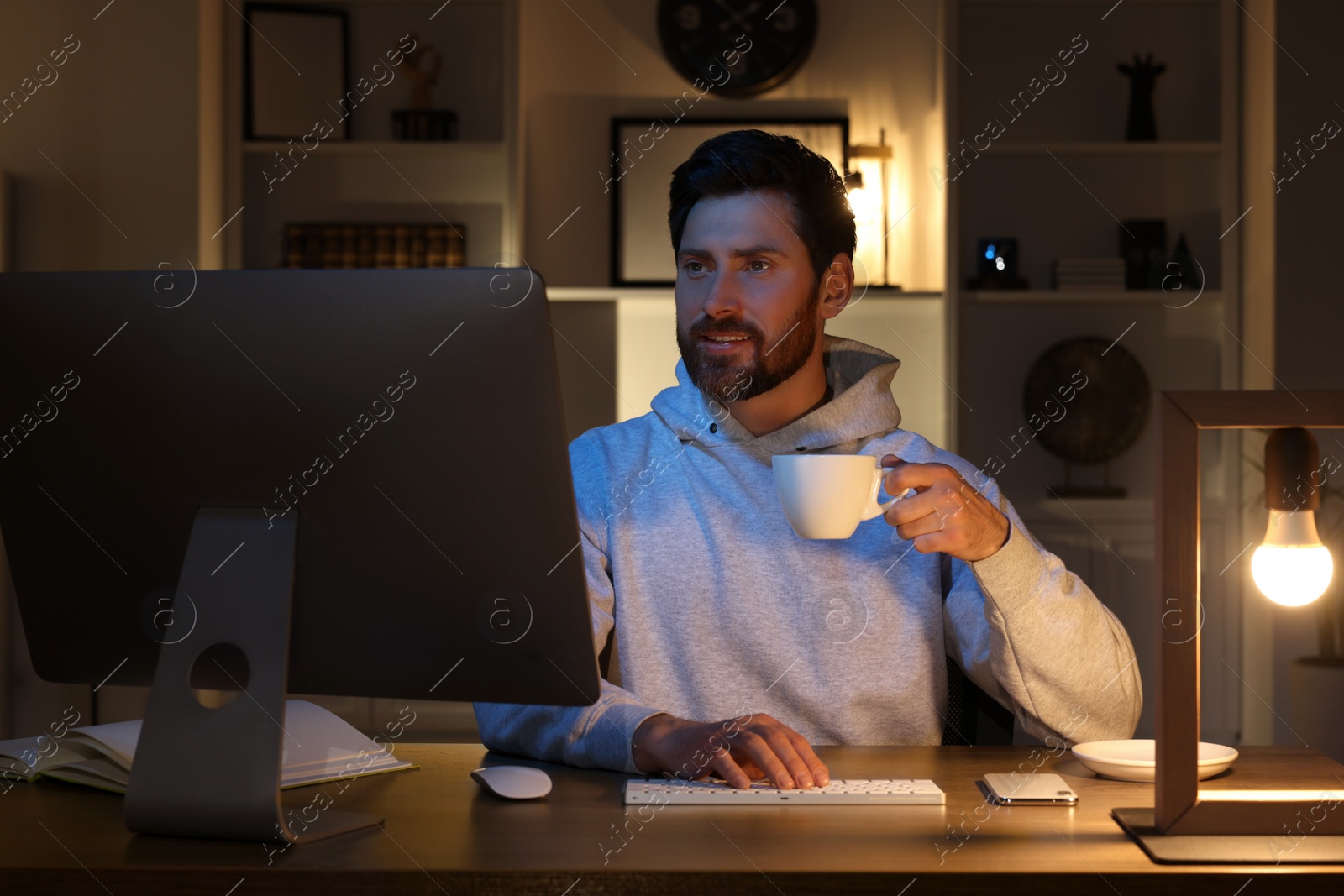 Photo of Home workplace. Man with cup of drink working with computer at wooden desk in room at night