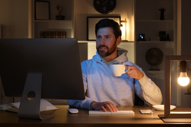 Photo of Home workplace. Man with cup of drink working with computer at wooden desk in room at night