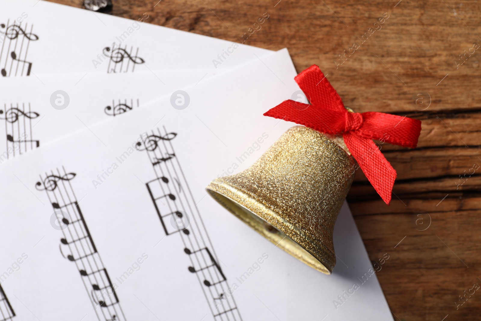 Photo of Golden shiny bell with red bow and music sheets on wooden table, flat lay. Christmas decoration