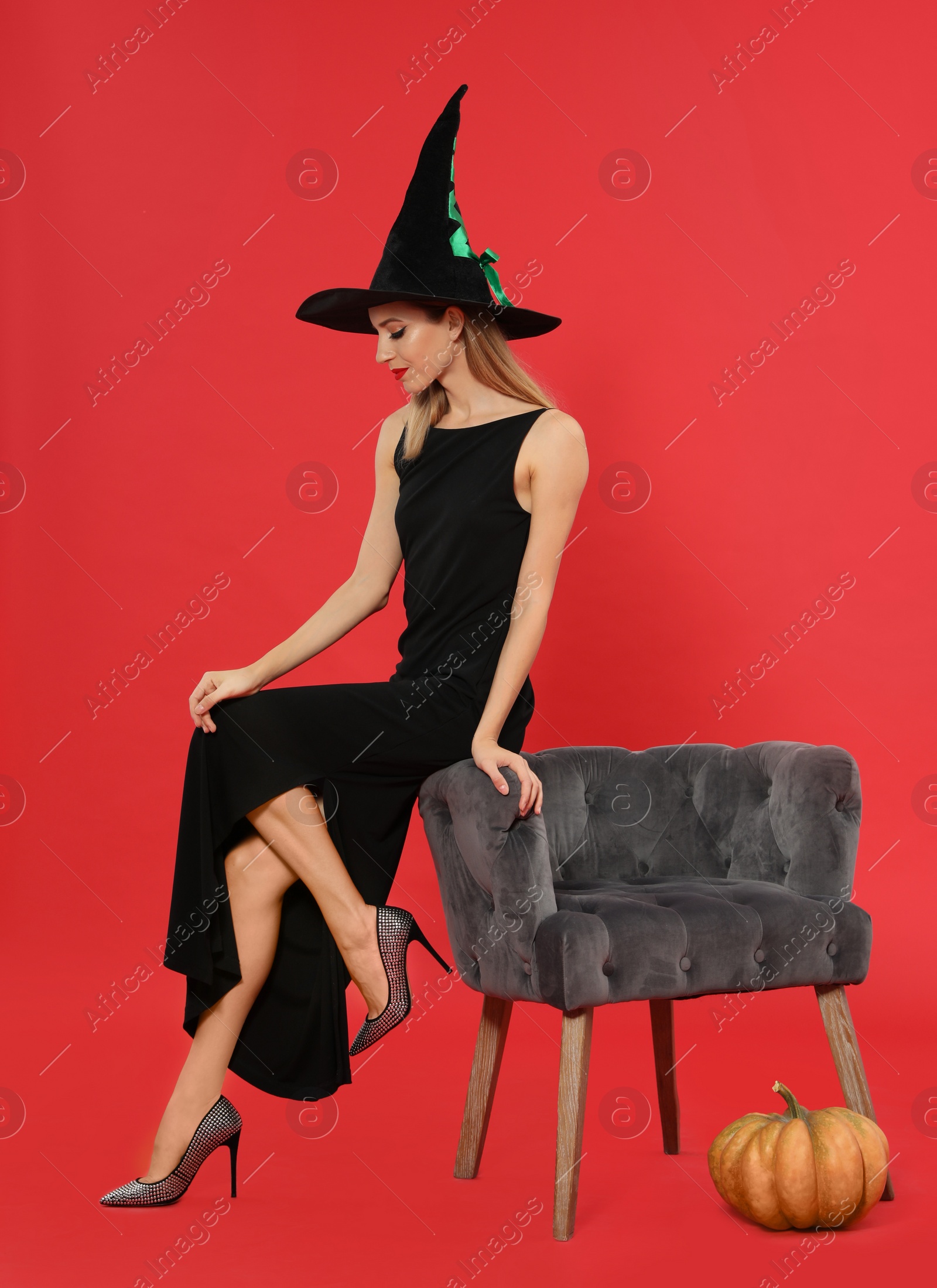 Photo of Beautiful woman wearing witch costume near armchair and pumpkin on red background. Halloween party