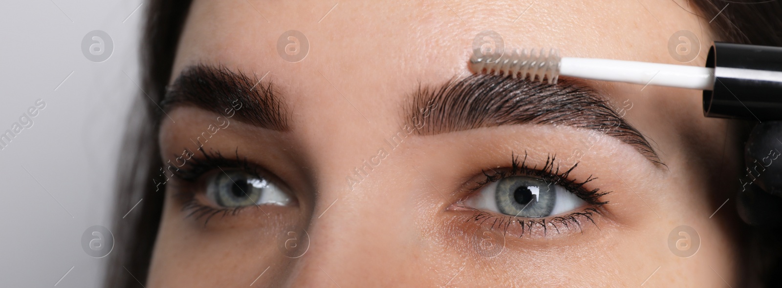 Photo of Beautician fixing woman's eyebrows with gel after tinting on light grey background, closeup