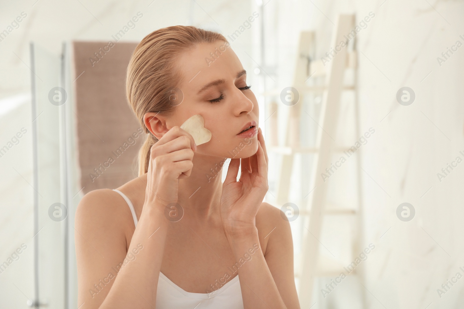 Photo of Beautiful young woman doing facial massage with gua sha tool in bathroom