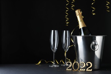 Photo of Happy New Year 2022! Bottle of sparkling wine in bucket and glasses on table against black background, space for text