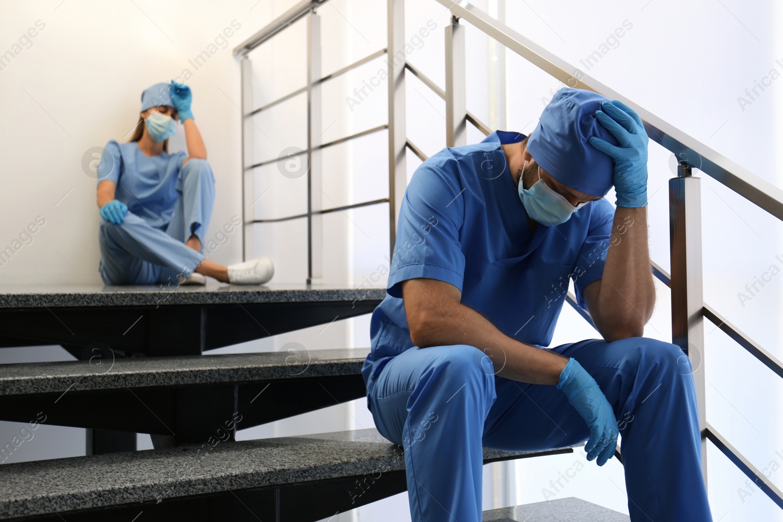 Photo of Exhausted doctors sitting on stairs indoors. Stress of health care workers during COVID-19 pandemic