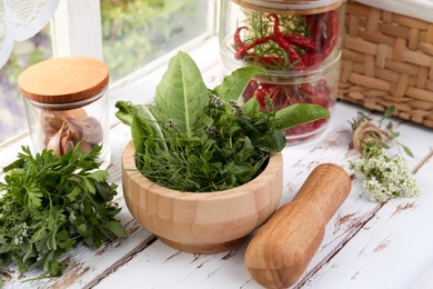 Mortar with pestle and fresh green herbs on white wooden table near window