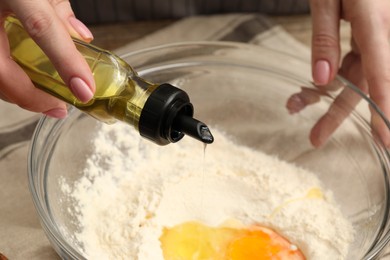 Woman pouring oil into bowl with flour and eggs at table, closeup