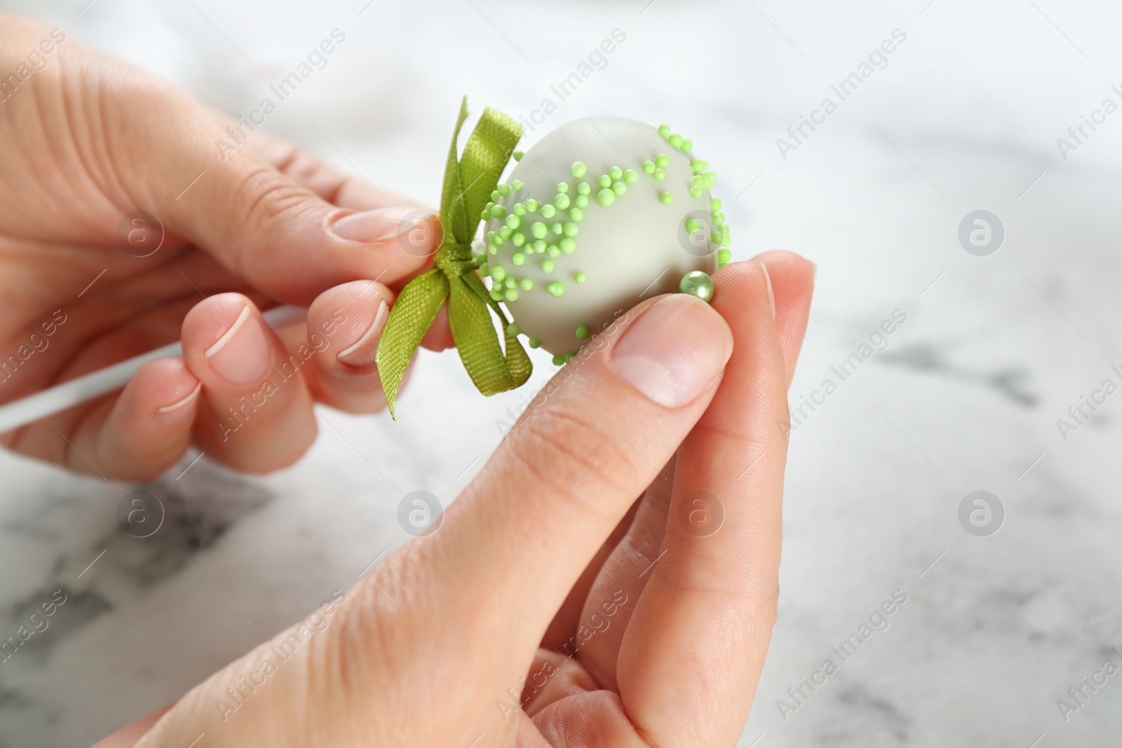 Photo of Young woman decorating tasty cake pop with green sprinkles at white marble table, closeup