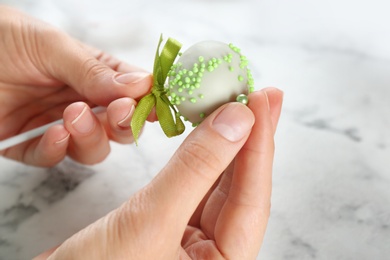 Young woman decorating tasty cake pop with green sprinkles at white marble table, closeup