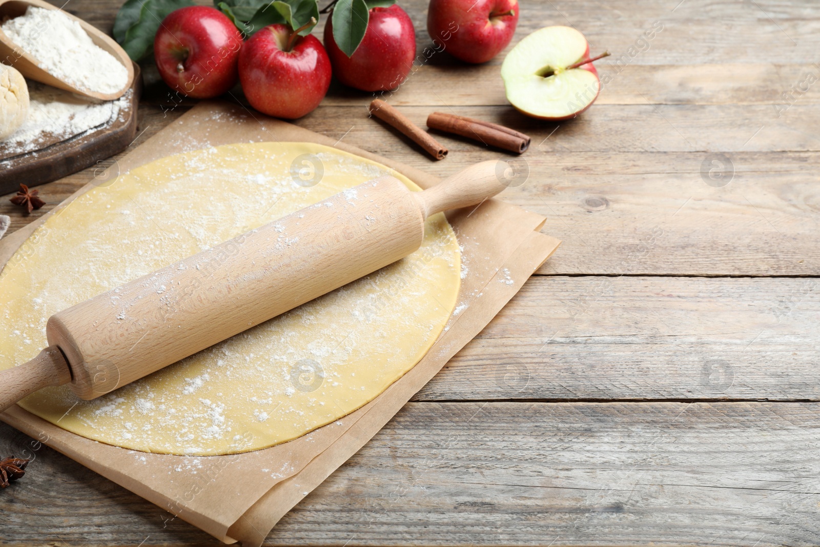 Photo of Rolling pin, raw dough and ingredients on wooden table, space for text. Baking apple pie