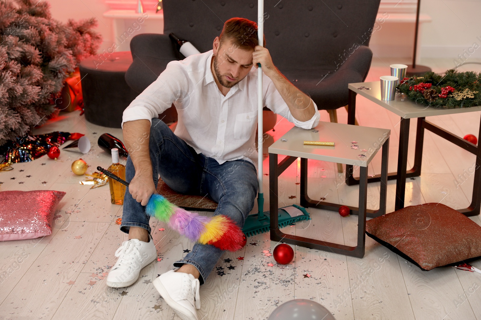 Photo of Man with dusting brush and mop suffering from hangover in messy room after New Year party