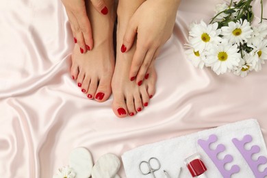 Photo of Woman with stylish red toenails after pedicure procedure, tools and chamomile flowers on light silk fabric, top view