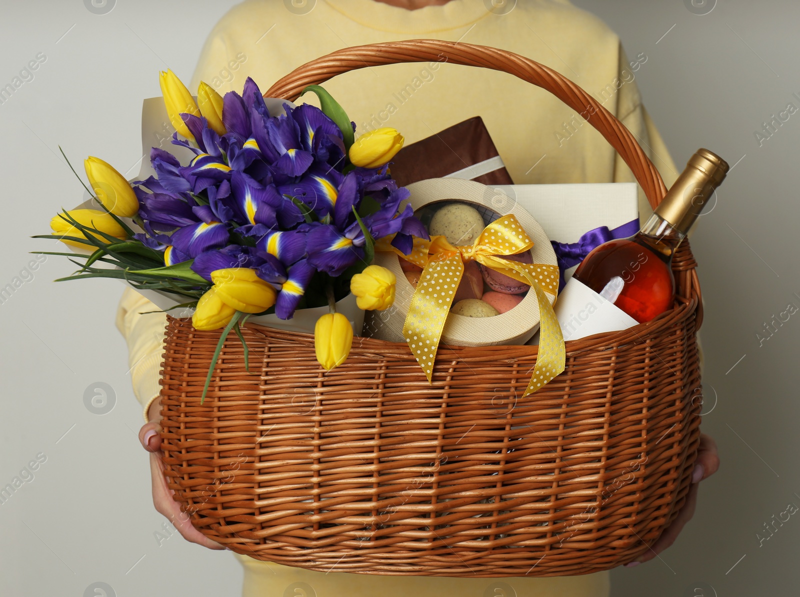 Photo of Woman holding wicker basket with gifts, bouquet and wine on grey background, closeup