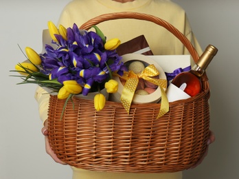 Woman holding wicker basket with gifts, bouquet and wine on grey background, closeup