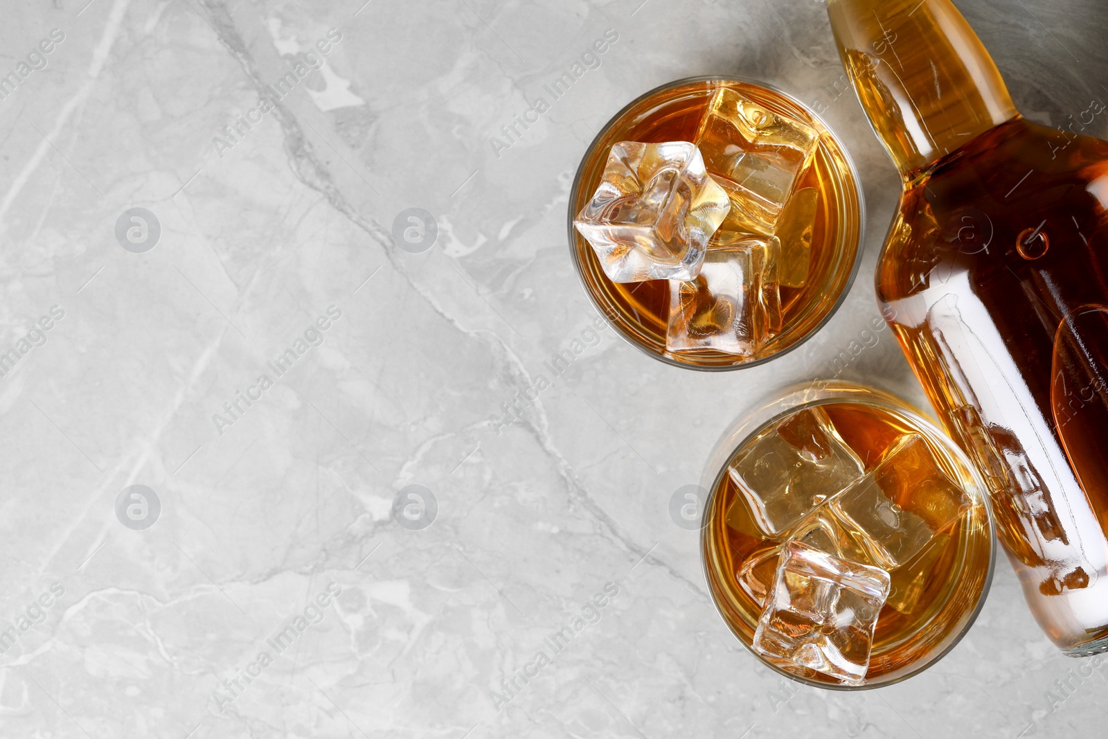 Photo of Whiskey with ice cubes in glasses and bottle on light grey textured table, flat lay. Space for text