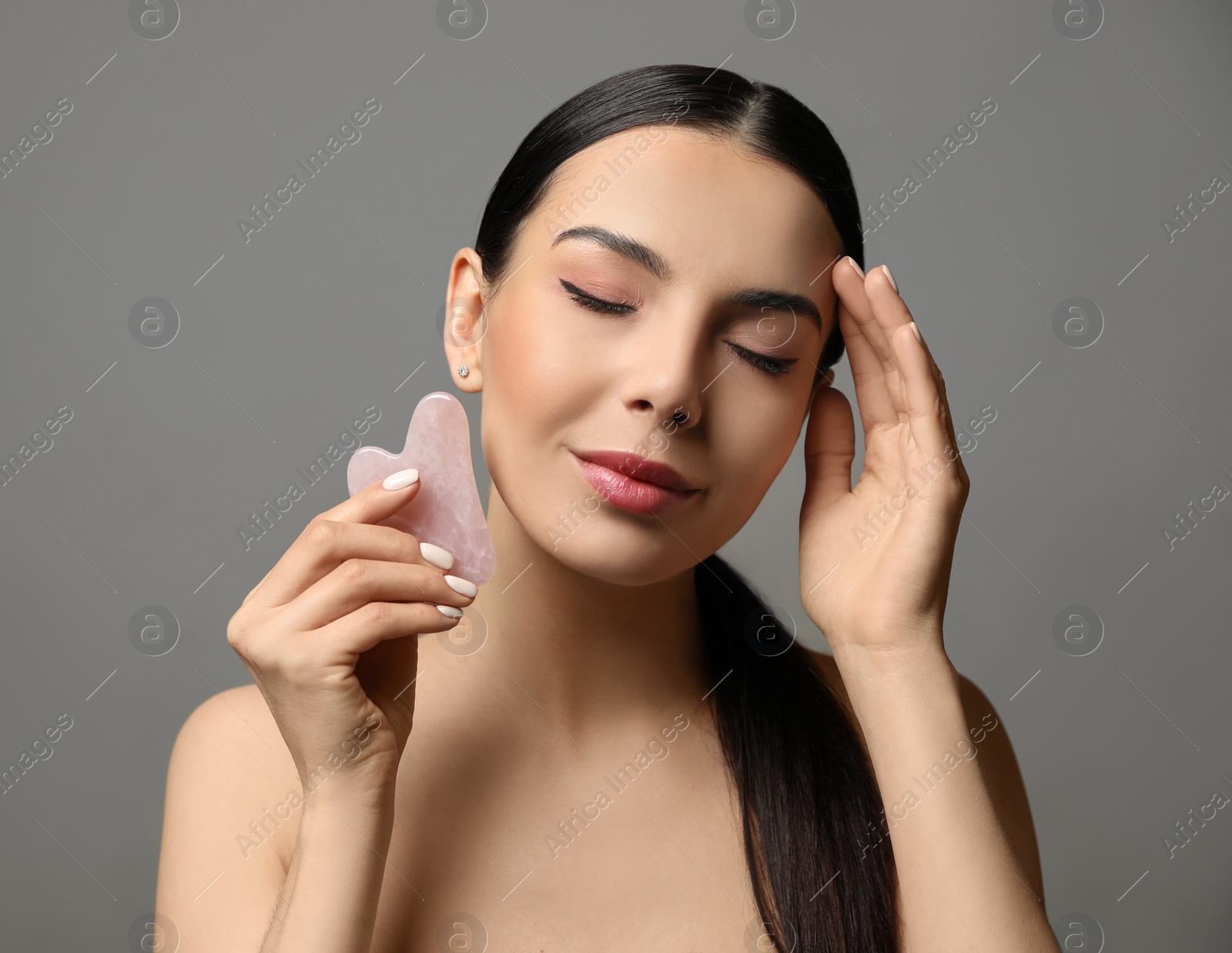 Photo of Beautiful young woman doing facial massage with gua sha tool on grey background