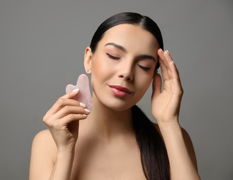 Beautiful young woman doing facial massage with gua sha tool on grey background