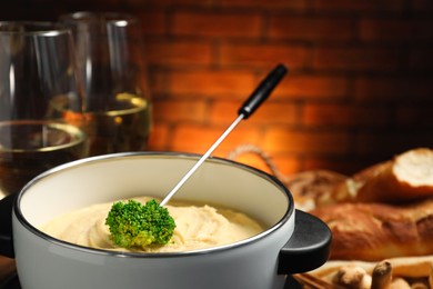 Fork with piece of broccoli, melted cheese in fondue pot, wine and snacks on blurred background, closeup