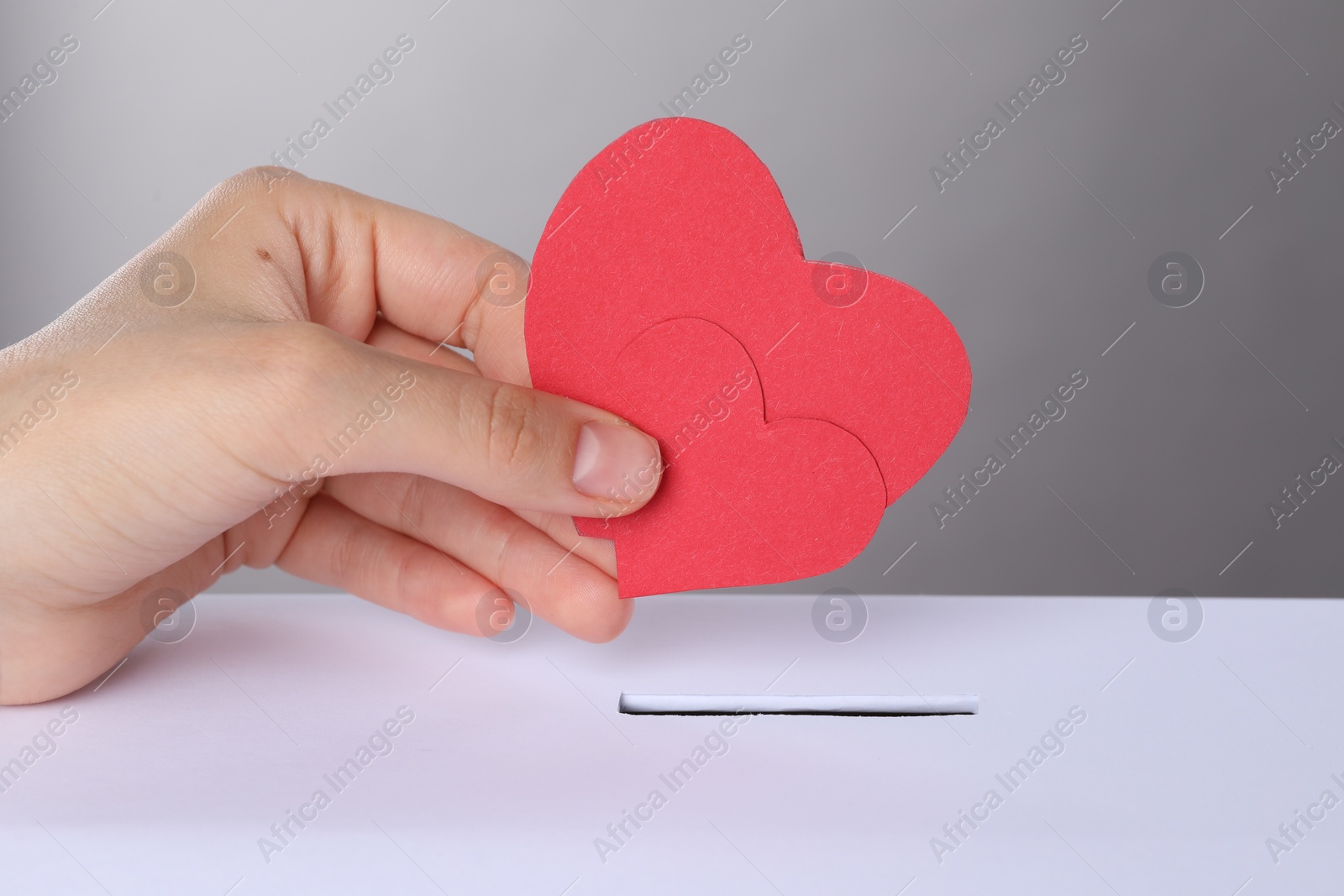 Photo of Woman putting red hearts into slot of donation box against grey background, closeup