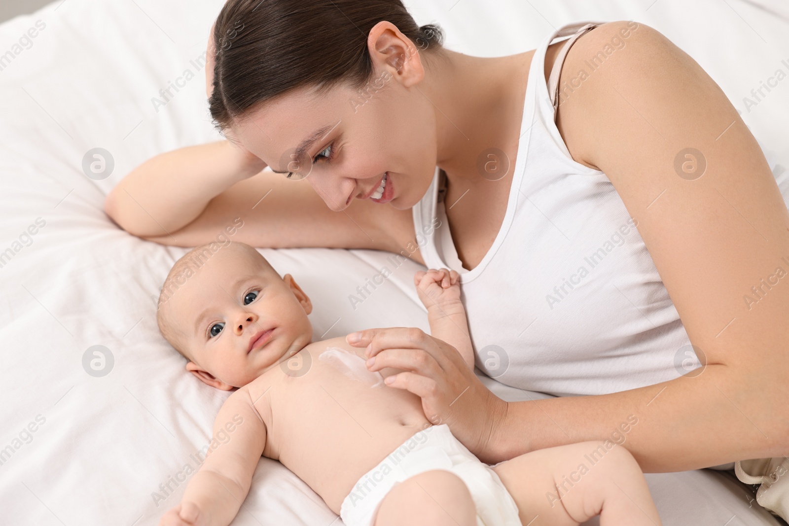 Photo of Happy young woman applying body cream onto baby`s skin on bed