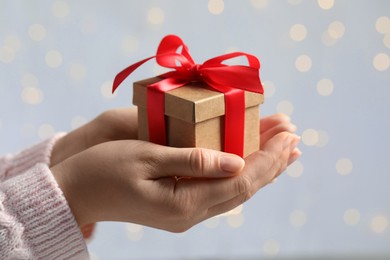 Woman holding gift box with red bow against blurred festive lights, closeup. Bokeh effect