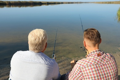 Father and adult son fishing together from riverside on sunny day