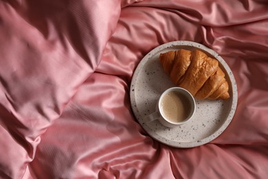 Photo of Tray with tasty croissant and cup of coffee on beautiful pink silk linens, top view