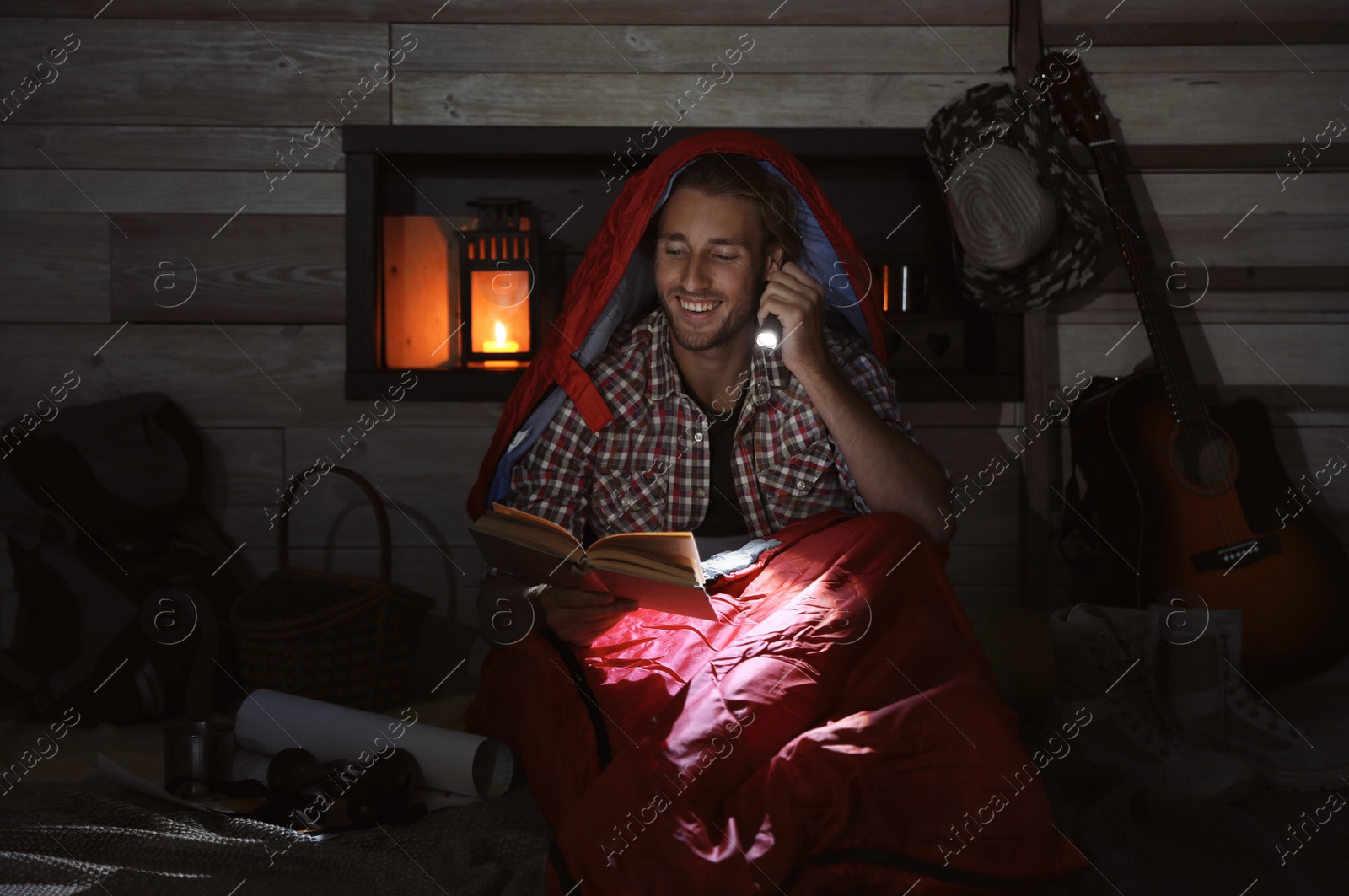 Photo of Young man in sleeping bag reading book with flashlight indoors