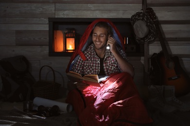 Photo of Young man in sleeping bag reading book with flashlight indoors