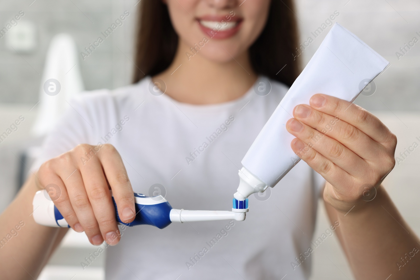 Photo of Woman squeezing toothpaste from tube onto electric toothbrush in bathroom, closeup