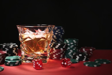 Photo of Casino chips, dice and glass of whiskey on red table against black background, space for text
