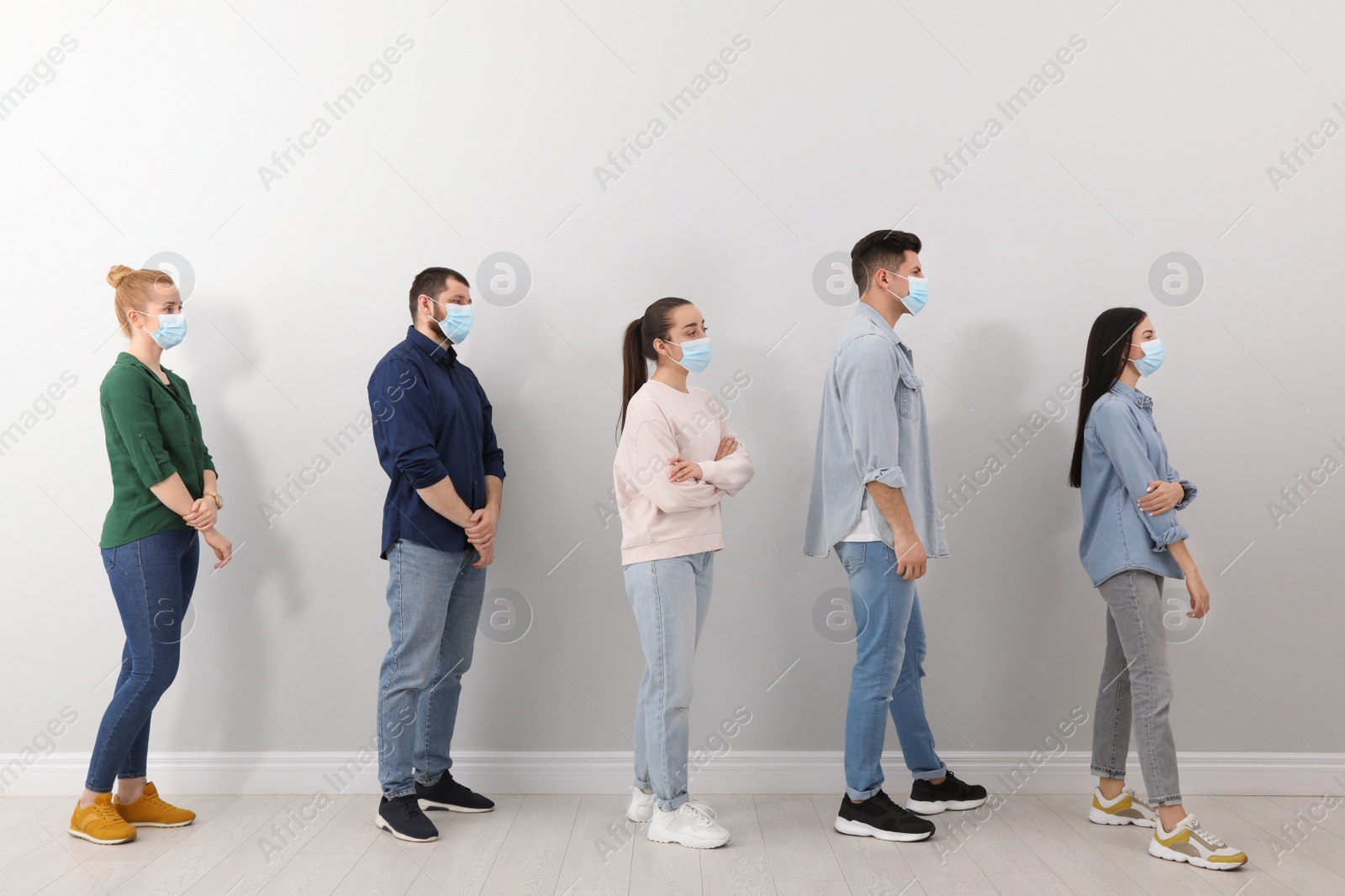 Photo of People with protective masks waiting in queue near light wall indoors. Social distancing during Covid-19 pandemic