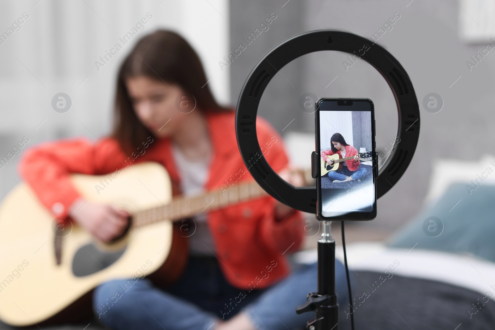 Photo of Teenage blogger playing guitar while streaming at home, focus on smartphone