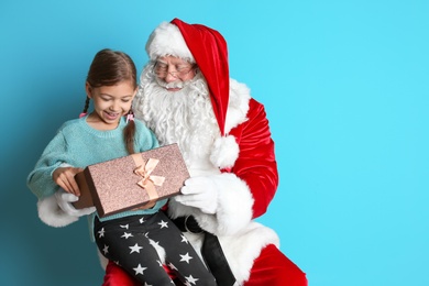 Little girl with gift box sitting on authentic Santa Claus' lap against color background