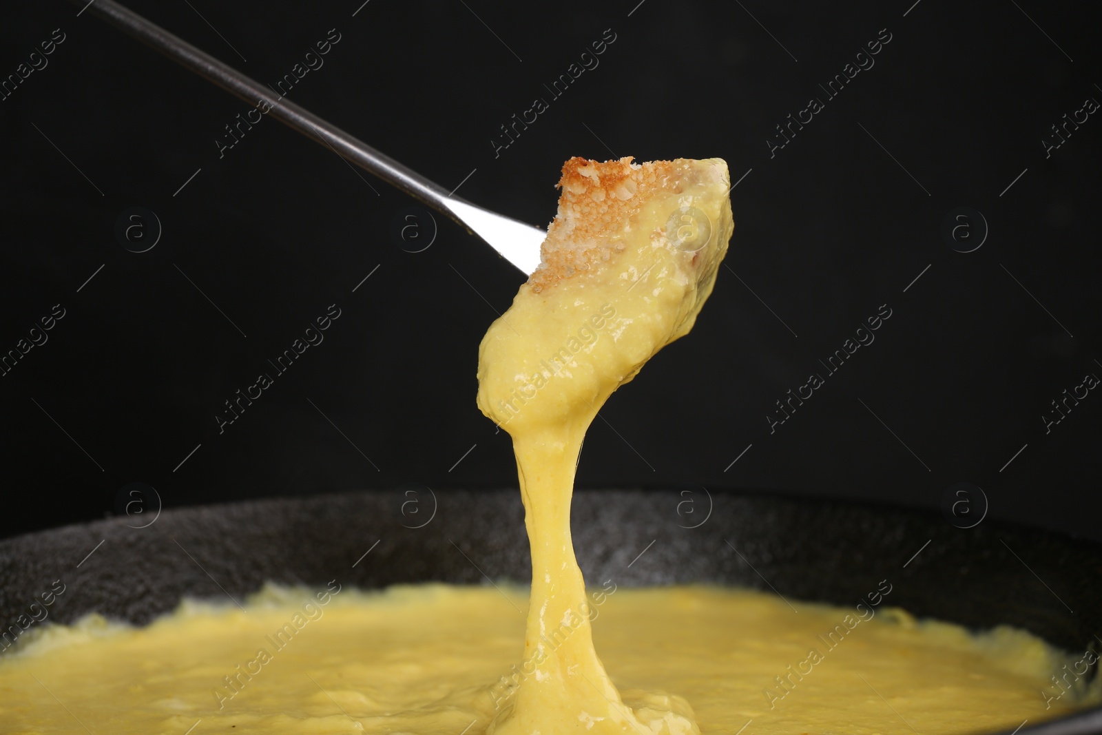 Photo of Dipping piece of bread into fondue pot with tasty melted cheese against dark gray background, closeup