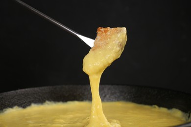 Dipping piece of bread into fondue pot with tasty melted cheese against dark gray background, closeup