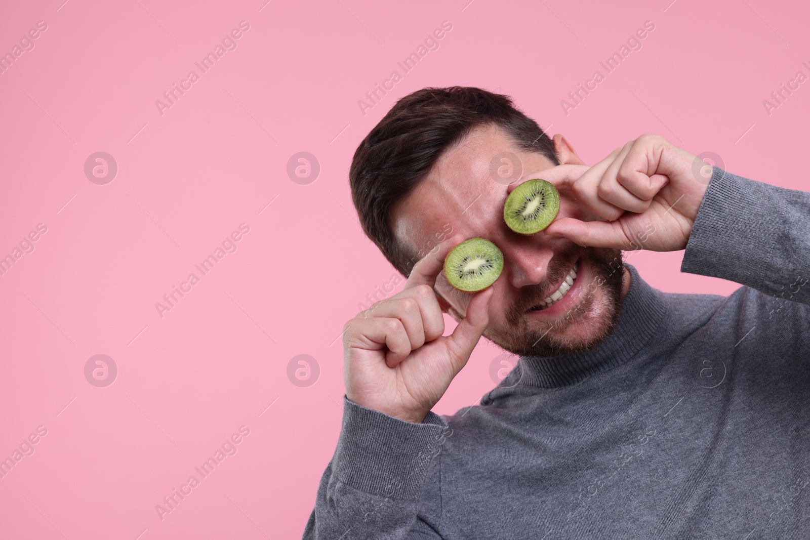 Photo of Man covering his eyes with halves of kiwi on pink background. Space for text