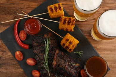 Glasses of beer, delicious grilled ribs, corn and sauces on wooden table, flat lay