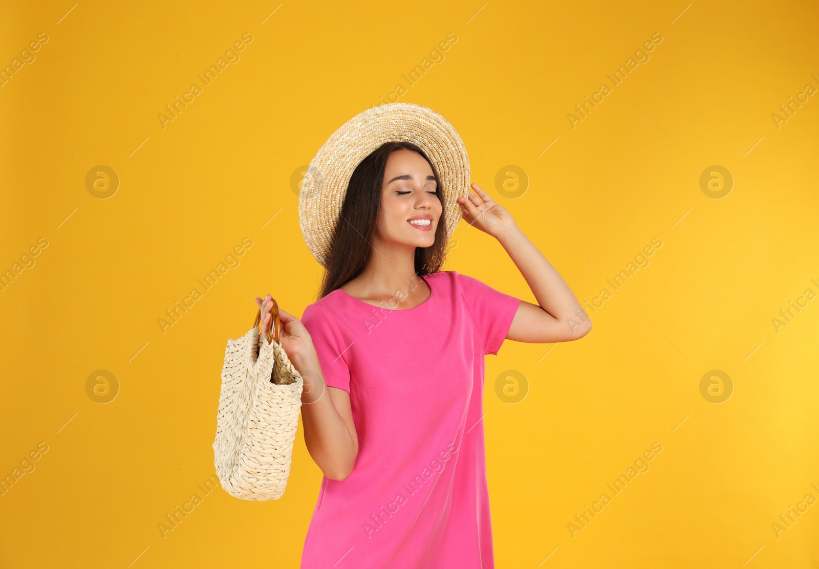 Photo of Young woman wearing stylish pink dress with straw bag on yellow background